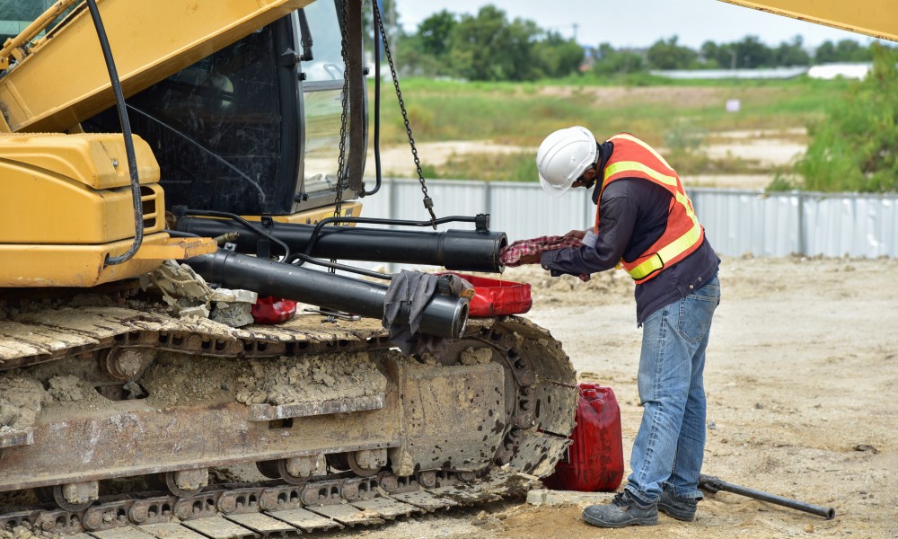 A heavy equipment mechanic handles repairs on the hydraulic system on a yellow excavator at a job site.