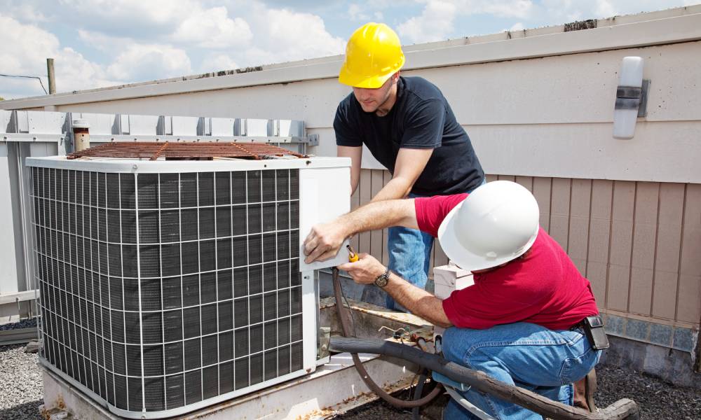 Two workers in differently colored hard hats work on an HVAC unit on top of a roof with sky and clouds in the background.