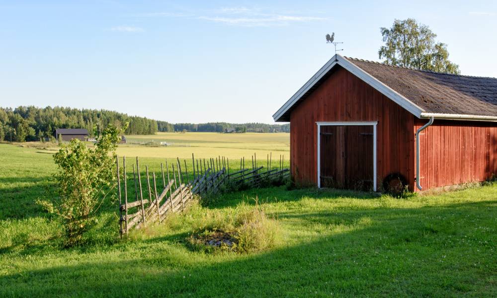 A small red barn stands on a vast piece of farmland. Next to the barn, a wooden fence looks like it is falling apart.