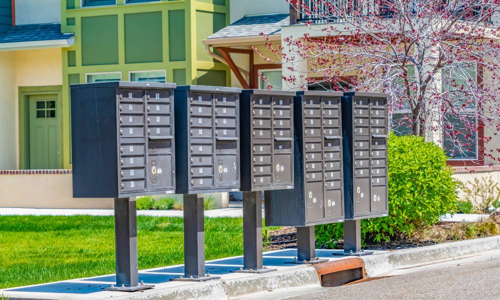 A row of grey cluster mailboxes sit on a sunny street surrounded by grass and backed by several homes.