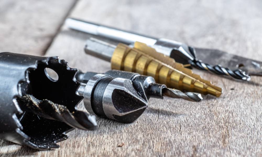 A set of drill bits lies on a gray wooden surface. Most of the bits are silver, while one has a gold color.