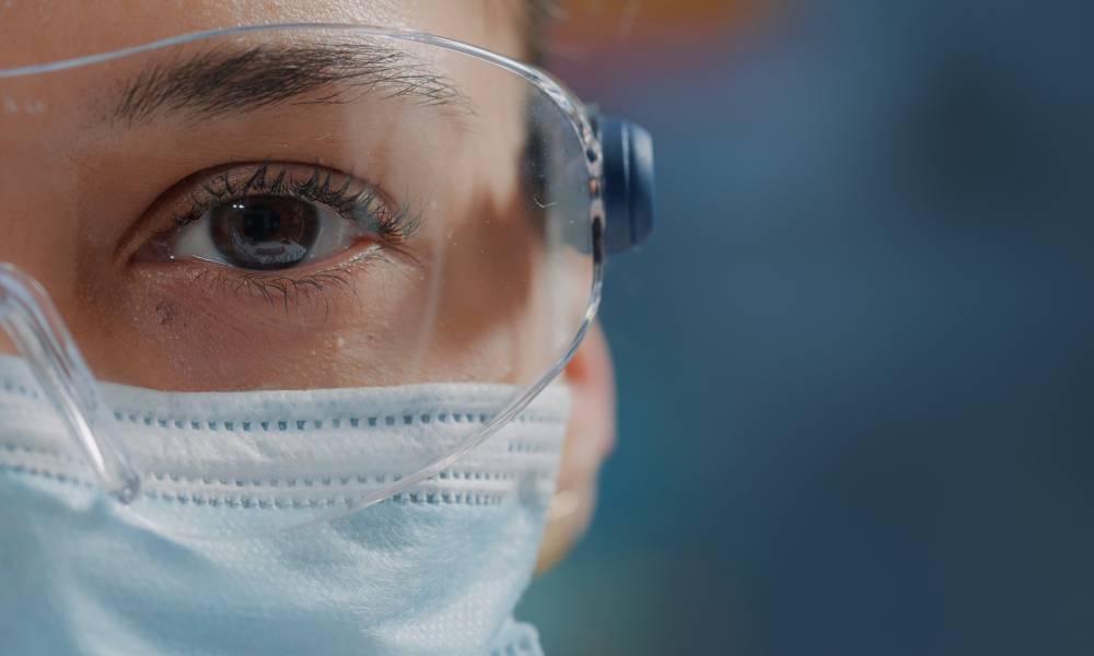 Close-up of a woman wearing safety glasses over her eyes and a face mask. The background behind her is blue.