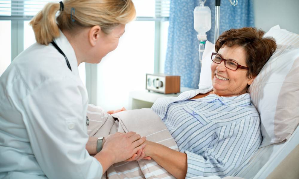 A doctor sits at the hospital bedside and holds her patient's hand. The patient lies in bed and smiles at the doctor.