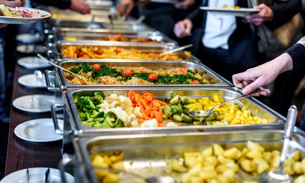 A line of food trays served buffet style. A hand grabs a large metal spoon in the tray full of vegetables.