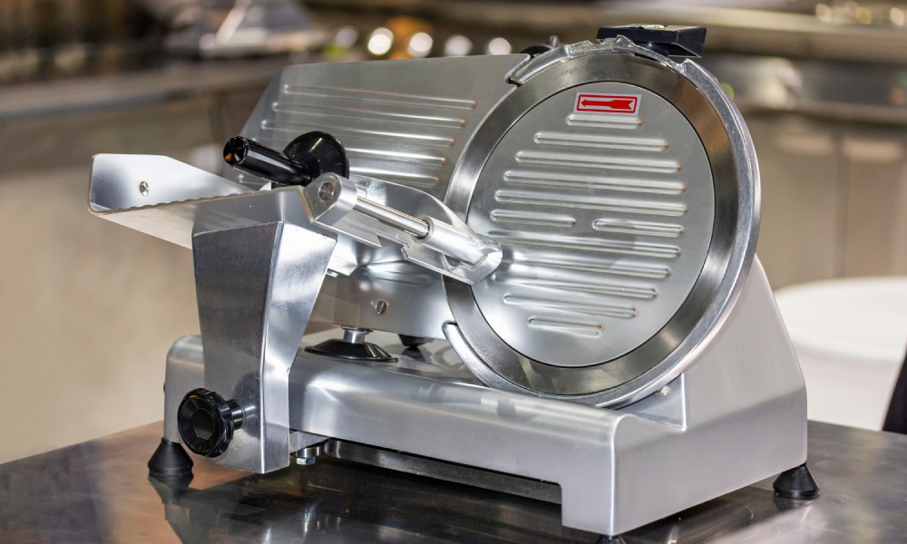 A modern semi-automatic meat slicer machine in an industrial kitchen setting on a stainless steel tabletop.