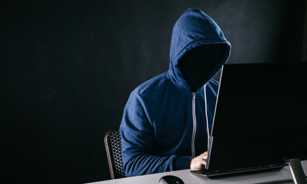 A person at a computer wearing a zip up hoodie over their head has their face completely blacked out with nothing behind them.